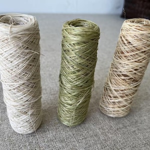 May include: Three spools of natural fiber yarn in varying shades. One is off-white, the second is a muted green, and the third is a light brown. The yarn is tightly wound, creating a cylindrical shape. Ideal for crafting and textile projects.