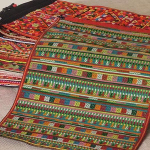 May include: A stack of colourful woven fabric strips with intricate patterns. The top strip is green, red, and blue with a geometric design. The strips are folded and stacked on a beige surface.