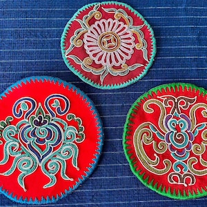 May include: Three round, embroidered coasters with intricate designs. Each coaster features a red base with detailed patterns in various colors, including blue, green, and gold. The edges are finished with a contrasting color.