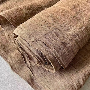 Könnte beinhalten: Eine Stoffrolle in Braun mit einer deutlich sichtbaren, strukturierten Webart. Das Material weist eine knittrige, unebene Oberfläche mit einem tiefen, erdigen Farbton auf. Der Stoff ist teilweise abgerollt und zeigt mehrere Lagen des Gewebes.