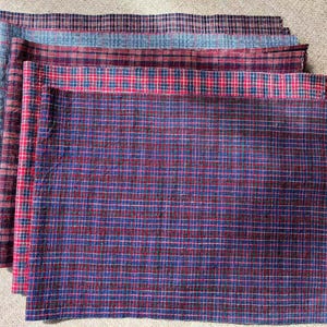 Könnte beinhalten: Ein Stapel karierter Stoffstücke in Blau-, Rot- und Burgundertönen. Der Stoff hat ein klassisches Karomuster mit unterschiedlich großen Karos. Die Stücke sind geschichtet und zeigen die verschiedenen Farbkombinationen und Texturen.