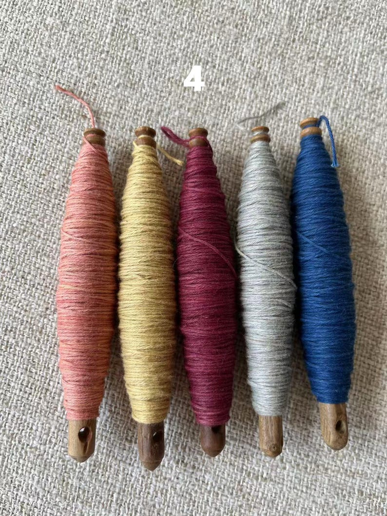 May include: Five colorful spools of thread in shades of pink, yellow, burgundy, gray, and blue. Each spool is wrapped around a wooden core with a small hole at the base. The number "4" is visible in the background.