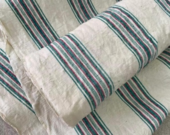 May include: A folded length of off-white fabric featuring a repeating pattern of vertical stripes in deep teal and thin red lines. The textile has a visible woven texture with small natural flecks throughout the light-colored base material.