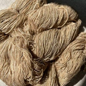 May include: A close-up of a pile of beige yarn. The yarn is soft and fluffy, and it has a subtle sheen.