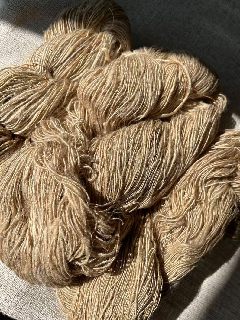 May include: A close-up of a pile of beige yarn. The yarn is soft and fluffy, and it has a subtle sheen.