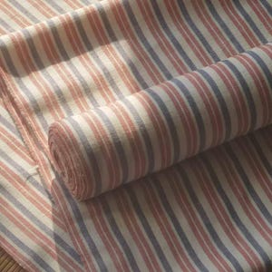 Op de afbeelding: Een rol witte stof met roze en blauwe strepen. De stof is dubbelgevouwen en de strepen lopen verticaal.