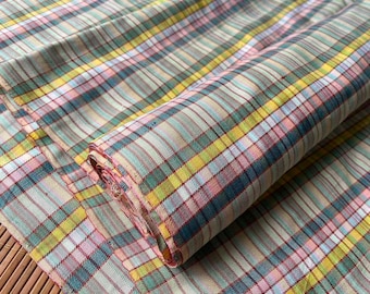 May include: A roll of fabric with a plaid pattern in shades of pink, yellow, teal, and gray. The fabric is folded and rolled, showing the pattern's repeating lines and colors. The fabric appears to be soft and lightweight.