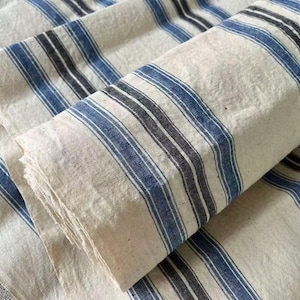 Pode incluir: Um rolo de tecido em tom off-white com um padrão repetitivo de listras verticais em tons de azul escuro e preto. O material possui uma textura tecida visível e está dobrado sobre si mesmo, exibindo o design listrado por toda a superfície.