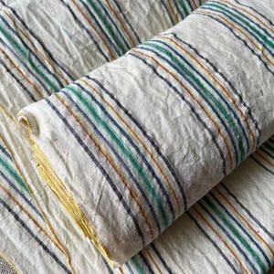 May include: A roll of off-white fabric featuring a repeating pattern of thin vertical stripes in shades of dark blue, emerald green, and mustard yellow. The textile has a visible textured weave and is folded to show multiple layers of the material.