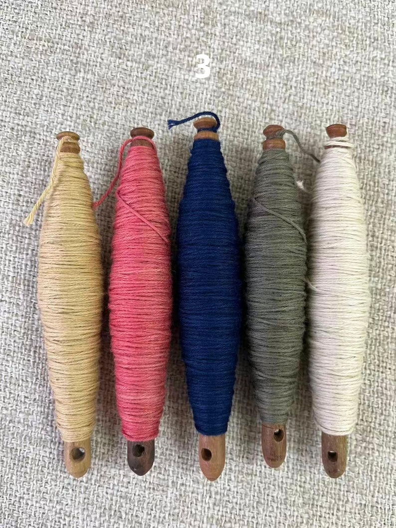 May include: Five colorful spools of thread. The spools are arranged in a row and are wrapped with thread in shades of beige, pink, navy blue, gray-green, and white. Each spool has a wooden base and top. The number "3" is visible in the background.