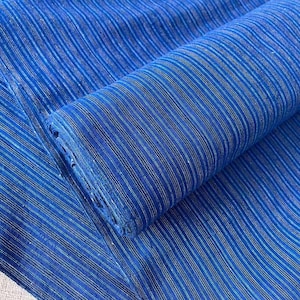 Könnte beinhalten: Ein Stoffballen mit einer leuchtend blauen Grundfarbe und dünnen, vertikalen Streifen in helleren Blau- und dezenten Gelbtönen. Das Textil weist eine durchgehend gleichmäßige Webstruktur auf und ist zu einer zylindrischen Form aufgerollt.