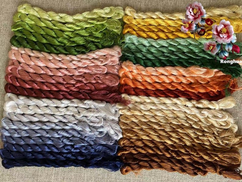 Pode incluir: Uma cole&ccedil;&atilde;o de carret&eacute;is de fio de bordado em v&aacute;rias cores, incluindo verde, rosa, vermelho, branco, azul e marrom. O fio est&aacute; disposto em filas, com cada fila contendo uma cor diferente. O fio &eacute; torcido e texturizado, criando uma exibi&ccedil;&atilde;o visualmente atraente.