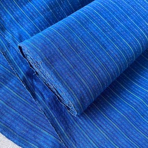 Puede incluir: Una pieza de tela azul intenso con un patrón de rayas verticales finas en tonos más claros de azul y verde. El tejido presenta una textura visible con pequeños guiones horizontales repetidos a lo largo de todo el diseño a rayas.