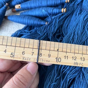 K&ouml;nnte beinhalten: Nahaufnahme eines Holzlineals, das ein B&uuml;ndel blaues Garn misst. Das Lineal ist mit Zahlen von 4 bis 13 und dem Text "Made in China" gekennzeichnet.