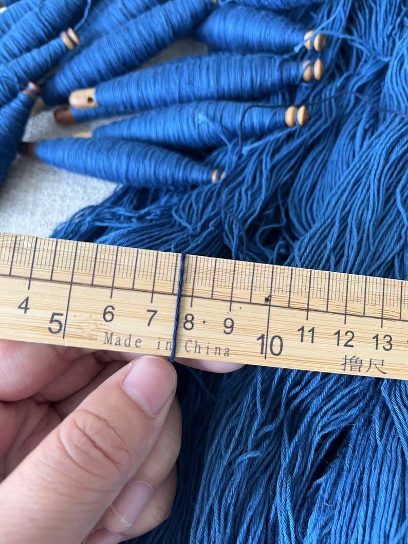 K&ouml;nnte beinhalten: Nahaufnahme eines Holzlineals, das ein B&uuml;ndel blaues Garn misst. Das Lineal ist mit Zahlen von 4 bis 13 und dem Text "Made in China" gekennzeichnet.