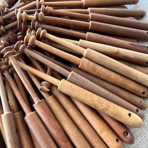 May include: A collection of wooden lace bobbins in various shades of brown. The bobbins have a cylindrical body with a rounded head and a pointed end. The wood grain is visible, and some bobbins have small holes.