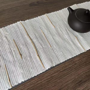 Puede incluir: Un camino de mesa de lino blanco con detalles de bambú tejido. El camino de mesa está sobre una superficie de madera oscura con una tetera negra.