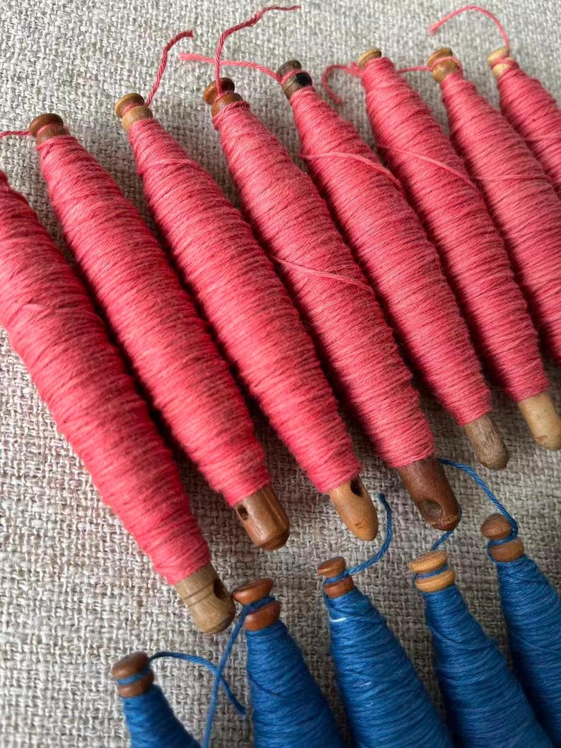 May include: Close-up of several pink thread spools with wooden bases, arranged diagonally. Below, a few blue thread spools are visible. The spools are likely for sewing or crafting, showcasing a variety of colors and textures.