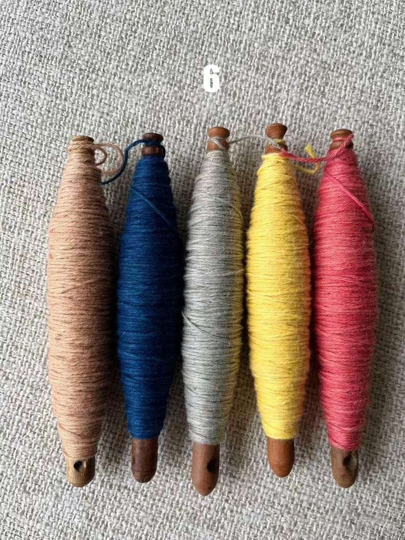 May include: Five wooden spools wrapped with colorful yarn in shades of brown, blue, gray, yellow, and red. The spools are arranged vertically, with the number "6" in white in the upper left corner. The yarn is tightly wound around the wooden spools.
