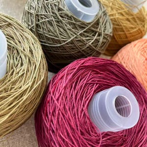May include: Close-up of several yarn balls in various colors, including olive green, gold, and deep pink. Each ball has a white plastic spool in the center. The yarn appears to be a fine weight, suitable for knitting or crochet projects.