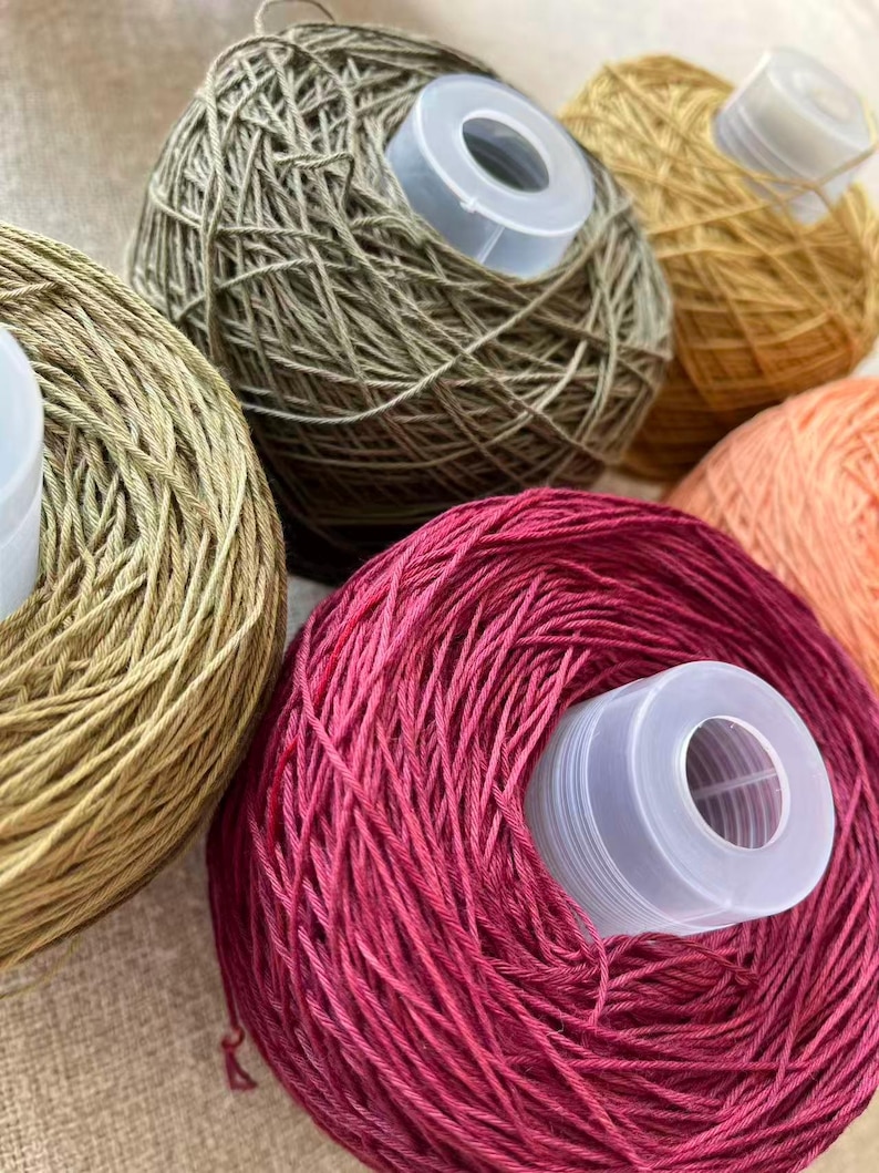 May include: Close-up of several yarn balls in various colors, including olive green, gold, and deep pink. Each ball has a white plastic spool in the center. The yarn appears to be a fine weight, suitable for knitting or crochet projects.