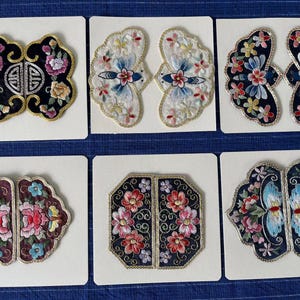 May include: A collection of six embroidered patches, each on a white card. The patches feature floral and geometric designs in various colors, including pink, blue, yellow, and black. The embroidery is detailed with gold thread accents.