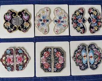 May include: A collection of six embroidered patches, each on a white card. The patches feature floral and geometric designs in various colors, including pink, blue, yellow, and black. The embroidery is detailed with gold thread accents.