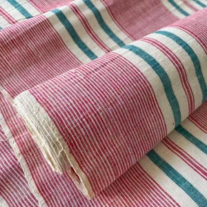 May include: A rolled-up textile with a striped pattern in red, white, and teal. The fabric is folded over, revealing the edge of the material. The stripes run vertically and are evenly spaced. The textile appears to be made of a natural fiber.