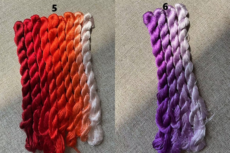 Pode incluir: Dois conjuntos de fio para bordar, um conjunto em tons de vermelho e laranja, o outro em tons de roxo e lavanda. O fio est&aacute; disposto em um padr&atilde;o de degrad&ecirc;, com os tons mais escuros na parte superior e os tons mais claros na parte inferior. O fio est&aacute; etiquetado com os n&uacute;meros 5 e 6.