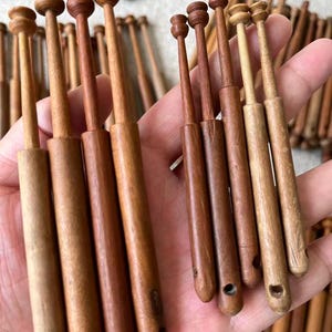 May include: A collection of wooden lace bobbins in various shades of brown. The bobbins have a rounded top and a tapered body, with a small hole at the bottom. Some bobbins are held in a hand, while others are scattered in the background.