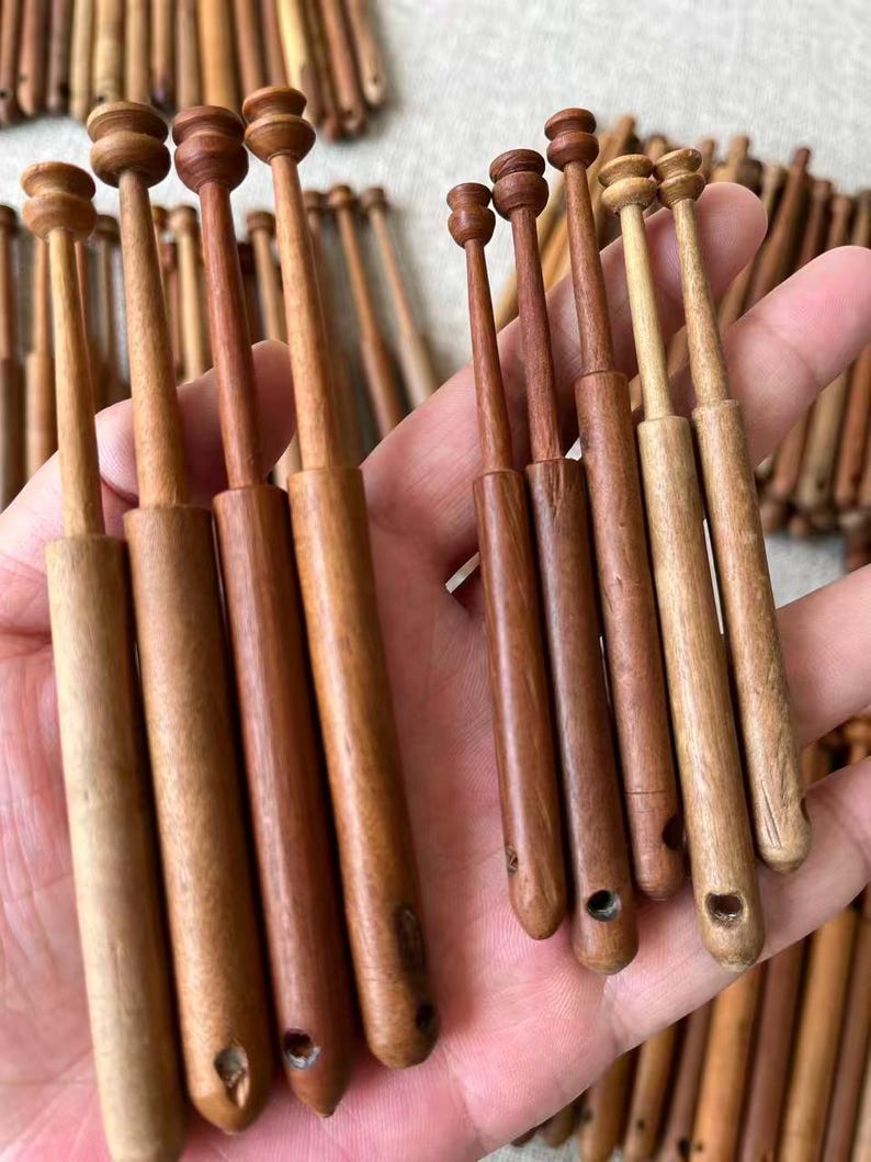 May include: A collection of wooden lace bobbins in various shades of brown. The bobbins have a rounded top and a tapered body, with a small hole at the bottom. Some bobbins are held in a hand, while others are scattered in the background.