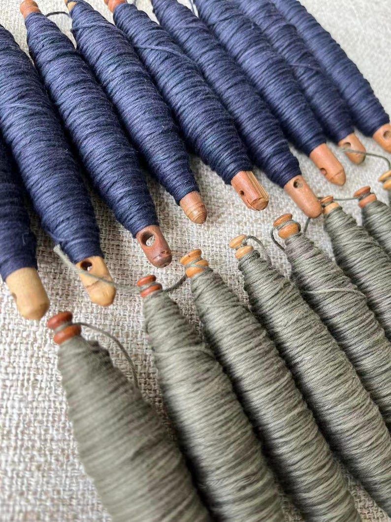 May include: An overhead shot of several spools of thread. The spools are navy blue and gray, with wooden bases. The navy blue spools are arranged in a row, while the gray spools are in a separate row. The background is a light beige fabric.