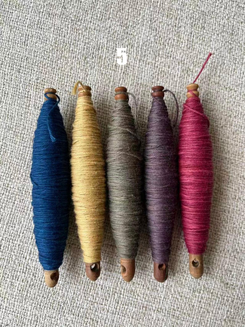 May include: Five spools of colorful yarn in shades of blue, yellow, green, purple, and red. Each spool is wrapped around a wooden core with a small hole at the bottom. The number "5" is printed in white at the top.