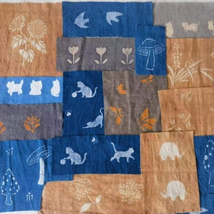 May include: A collection of fabric squares with various hand-painted designs, including mushrooms, cats, butterflies, and flowers. The fabric is a mix of blue, brown, and grey.