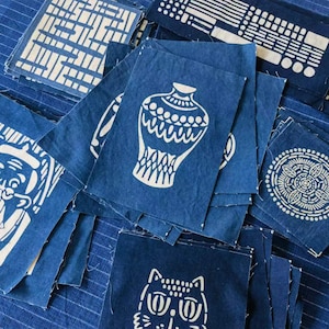 Puede incluir: Una colección de cuadrados de tela azul con diseños blancos. Los diseños incluyen un jarrón, una criatura y un patrón geométrico. Los cuadrados de tela están apilados uno encima del otro.