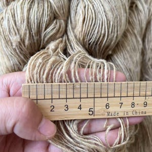 May include: A close-up of a skein of beige yarn with a wooden ruler measuring 5 inches. The yarn is soft and fluffy, with a subtle sheen.