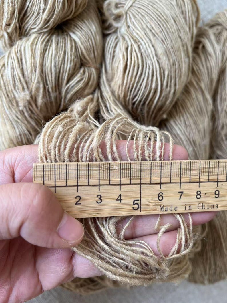May include: A close-up of a skein of beige yarn with a wooden ruler measuring 5 inches. The yarn is soft and fluffy, with a subtle sheen.