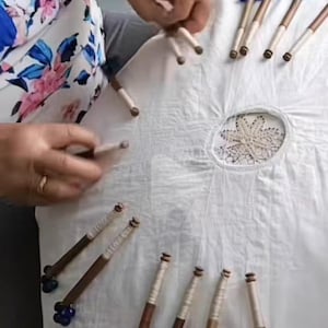 May include: A collection of wooden lace bobbins with blue glass bead ends, arranged on a white cloth. The bobbins have a dark brown wooden shaft and are partially wrapped with white thread. The cloth has a central embroidered design.