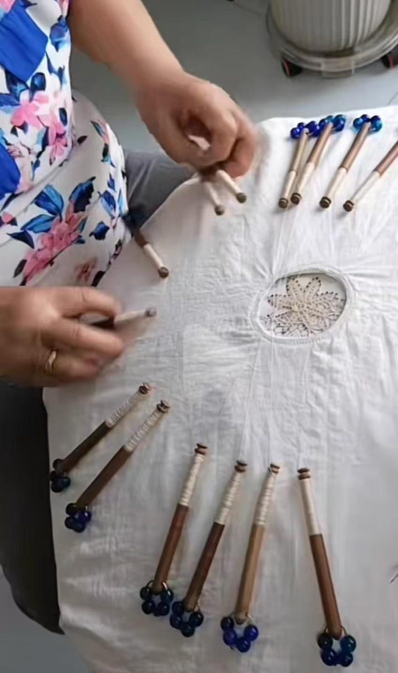 May include: A collection of wooden lace bobbins with blue glass bead ends, arranged on a white cloth. The bobbins have a dark brown wooden shaft and are partially wrapped with white thread. The cloth has a central embroidered design.
