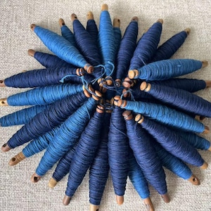 Puede incluir: Una colección de carretes de hilo azul e índigo, enrollados alrededor de bobinas de madera. Los carretes están dispuestos en un patrón circular, con las bobinas mirando hacia afuera.