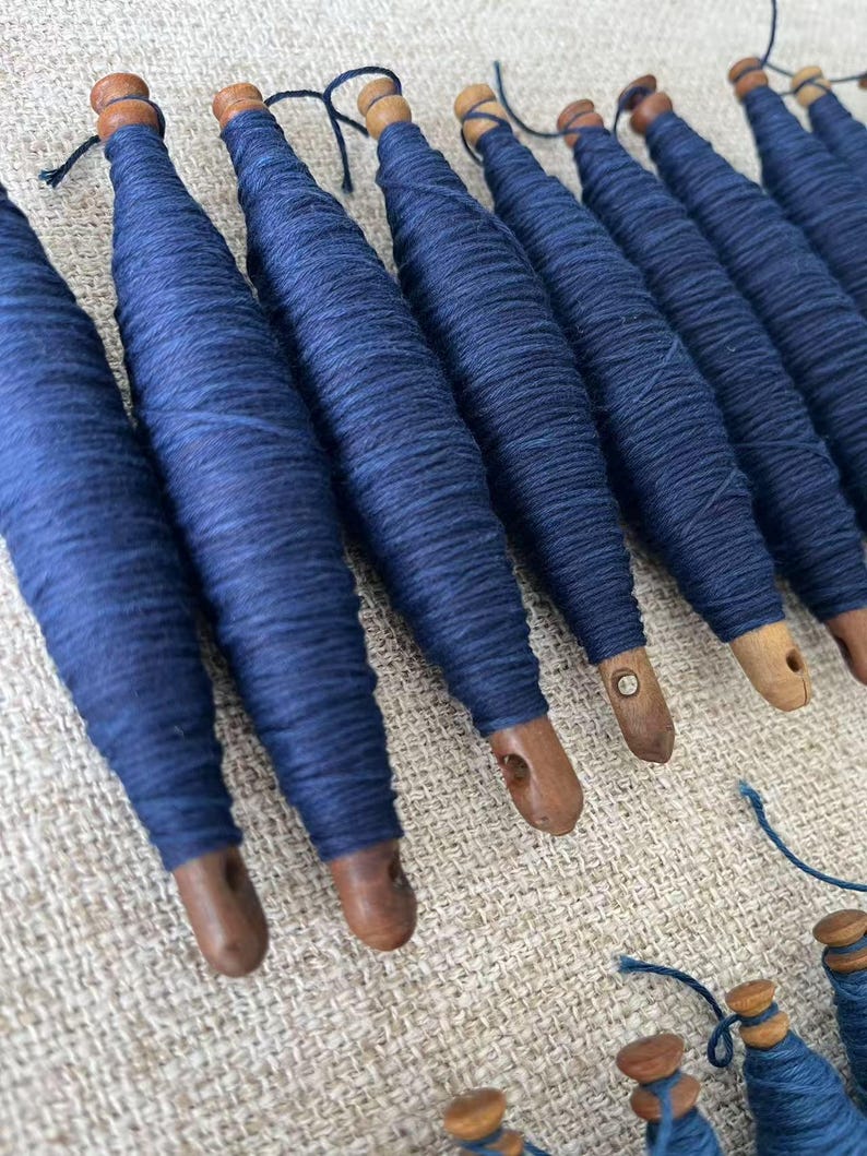 K&ouml;nnte beinhalten: Nahaufnahme einer Sammlung von Holzspulen, die mit dunkelblauem Garn gewickelt sind. Die Spulen sind in einer Reihe auf einer strukturierten Oberfl&auml;che angeordnet.