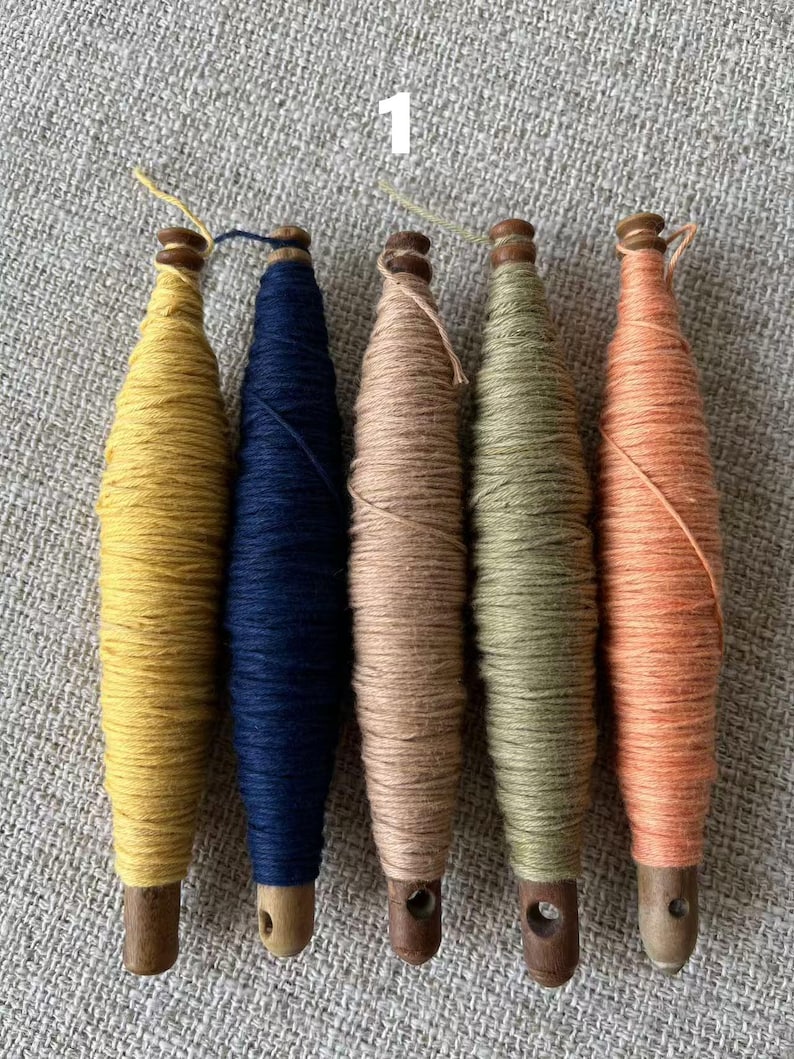 May include: Five colorful yarn spools in shades of yellow, navy blue, tan, olive green, and peach. Each spool is wound around a wooden base with a small hole at the bottom. The spools are arranged in a row on a textured gray surface.
