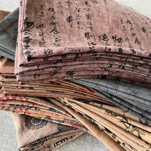 May include: A stack of folded fabric pieces in various colors, including muted pink, gray, and beige. The fabric is printed with black calligraphy and floral patterns. The textiles appear to be stacked on top of each other.