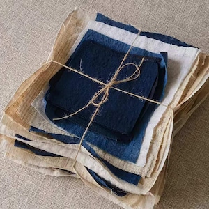 May include: A stack of fabric squares in various shades of blue and white, tied together with twine. The fabric is a natural linen or cotton blend, and the squares are layered to create a textured effect.