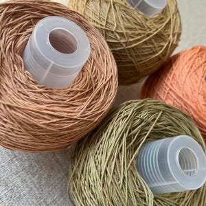 May include: Close-up of several yarn balls in shades of brown, green, and orange. Each ball has a white plastic spool in the center. The yarn appears to be a fine weight, suitable for knitting or crochet projects.