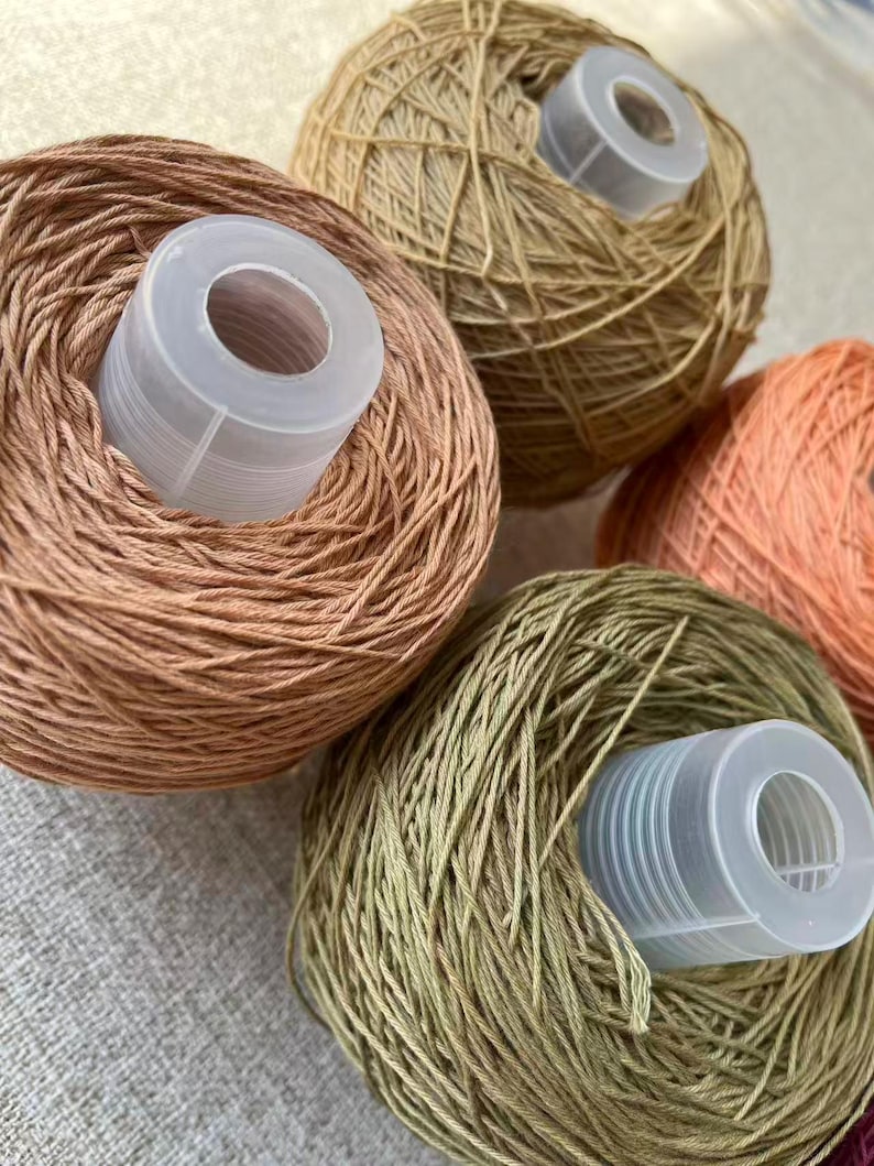 May include: Close-up of several yarn balls in shades of brown, green, and orange. Each ball has a white plastic spool in the center. The yarn appears to be a fine weight, suitable for knitting or crochet projects.