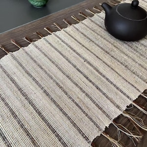 Peut inclure: Un napperon tissé beige et brun avec des bords frangés. Le napperon est fait de fibres naturelles et présente un motif rayé subtil.