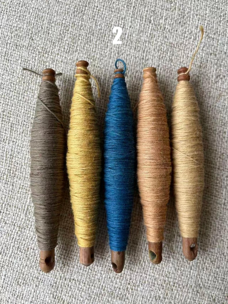 May include: Five wooden spools wrapped with colorful yarn. The spools are in shades of brown, yellow, blue, and tan. The spools are arranged in a row on a textured, light gray surface. The number "2" is visible in the background.