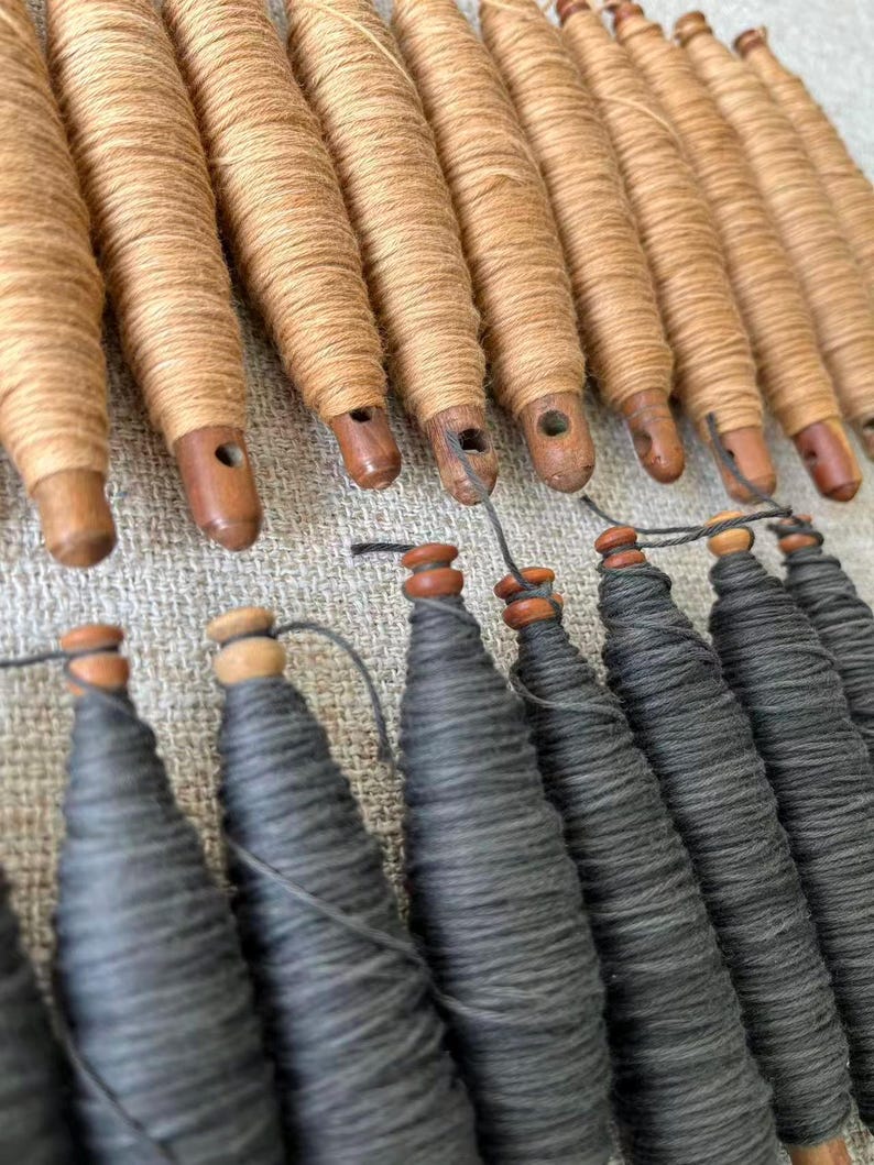 May include: A collection of wooden spools, some wrapped with tan thread and others with gray thread. The spools have a tapered shape and are arranged in rows, suggesting a textile or craft theme.