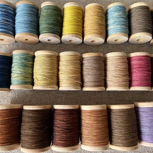 Puede incluir: Una colección de 18 carretes de hilo en varios colores, incluyendo azul, verde, amarillo, marrón, rojo y morado. Los carretes están dispuestos en dos filas, con nueve carretes en cada fila.